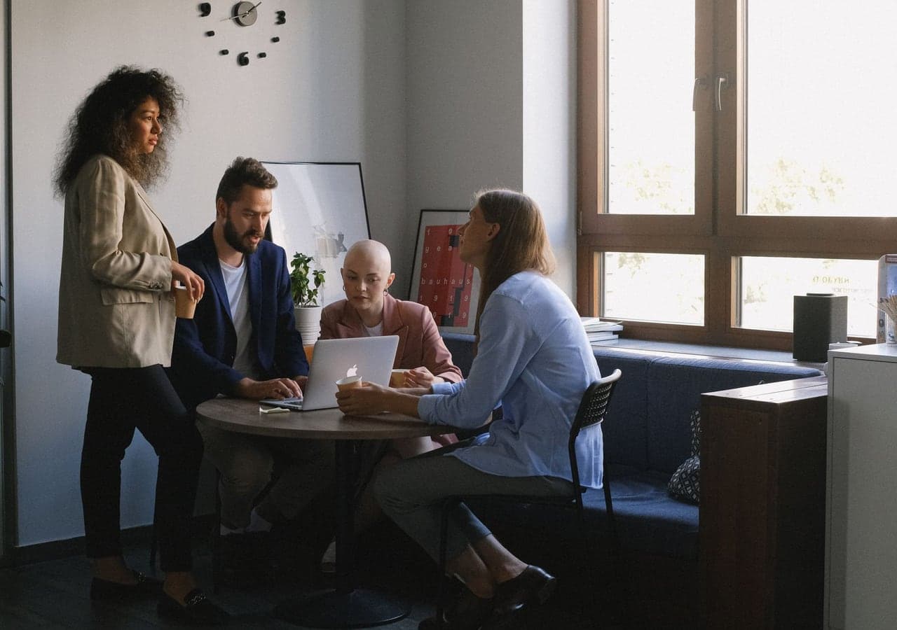 Four colleagues talking around a laptop at a table in a bright office