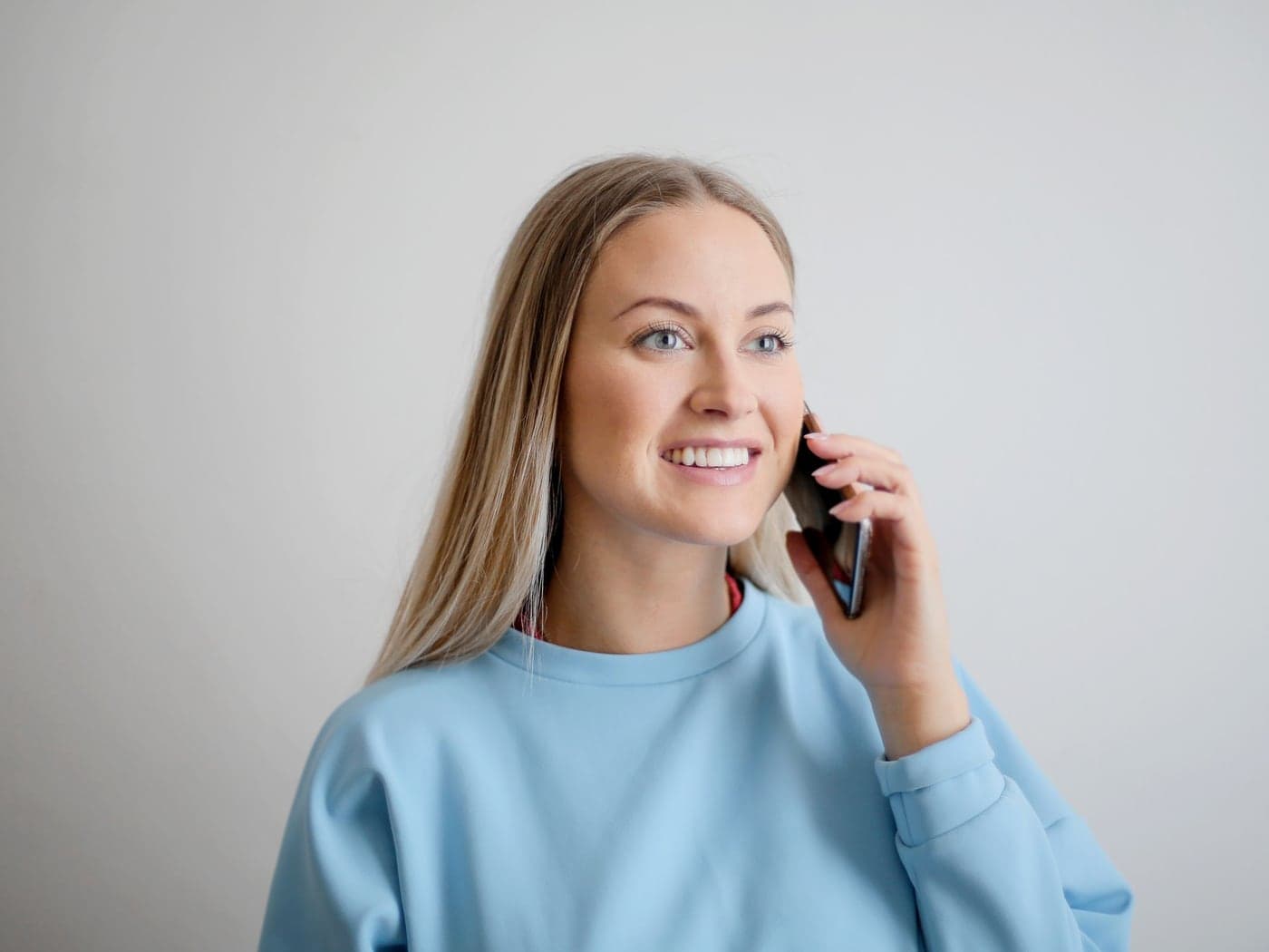 A smiling woman in a light blue sweatshirt talking on her phone