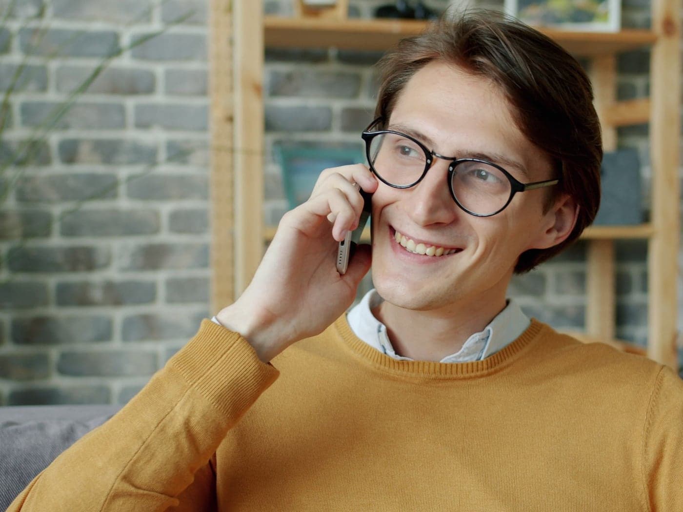 A smiling man in glasses and a mustard sweater on a phone call at home
