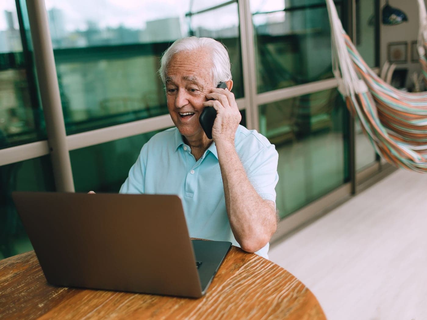 An older man smiling on a phone call at a laptop by the window